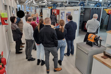 Eine Gruppe aus Männern und Frauen steht in einer modernen Werkhalle mit Lüftungssystemen und mehreren Arbeitskabinen; sie lauschen einer Frau, die vor einer Infotafel spricht, während im Vordergrund Werkbänke und Maschinen zu sehen sind.