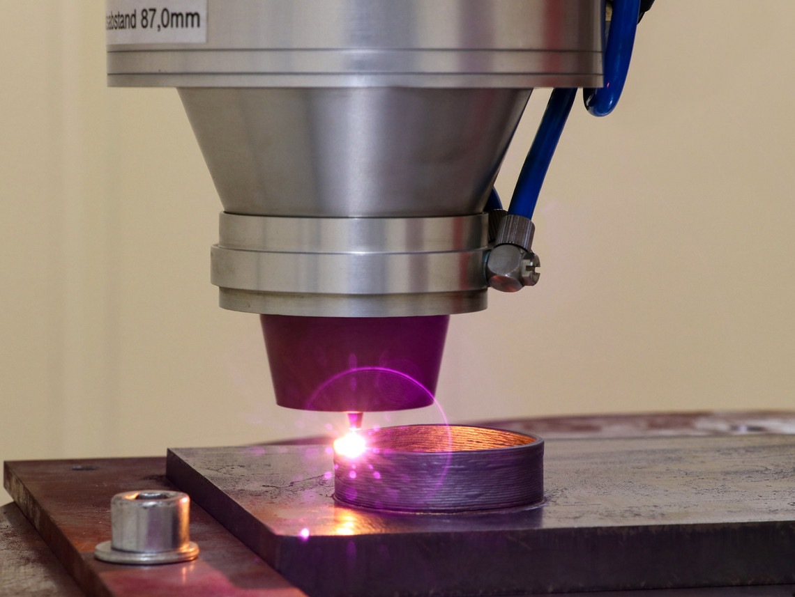 Ein Laser-Bearbeitungskopf schmilzt punktgenau Metallpulver auf ein Werkstück, wodurch schichtweise ein zylindrischer Aufbau entsteht; die violette Laserstrahlung ist deutlich sichtbar.