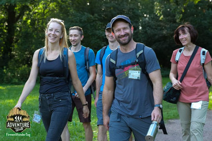 Sechs gut gelaunte Erwachsene mit Namensschildern und Rucksäcken wandern auf einem Waldweg bei Sonnenschein, unten links ist das Logo des „Adventure Walk FirmenWanderTag“ eingeblendet.