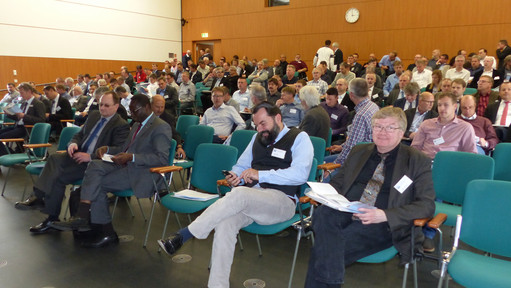 Ein vollbesetzter Vortragssaal mit überwiegend männlichem Publikum vor Beginn einer Veranstaltung, viele Personen halten Tagungsunterlagen in der Hand. In der ersten Reihe sitzt Dr. Karamba Diaby, Mitglied des Deutschen Bundestages 
