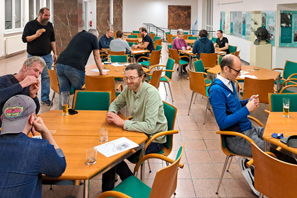 Mehrere Personen sitzen in einem Raum an Holztischen und spielen Karten, lebhafte Atmosphäre.