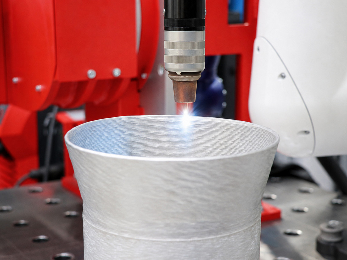 Ein Bearbeitungskopf erzeugt einen hellen Lichtpunkt auf der Oberkante eines zylindrischen Metallbauteils mit sichtbar geschichteter Oberflächenstruktur. Das Bauteil steht auf einem Arbeitstisch mit Lochraster, im Hintergrund sind Teile einer roten und weißen Maschinenanlage zu sehen.
