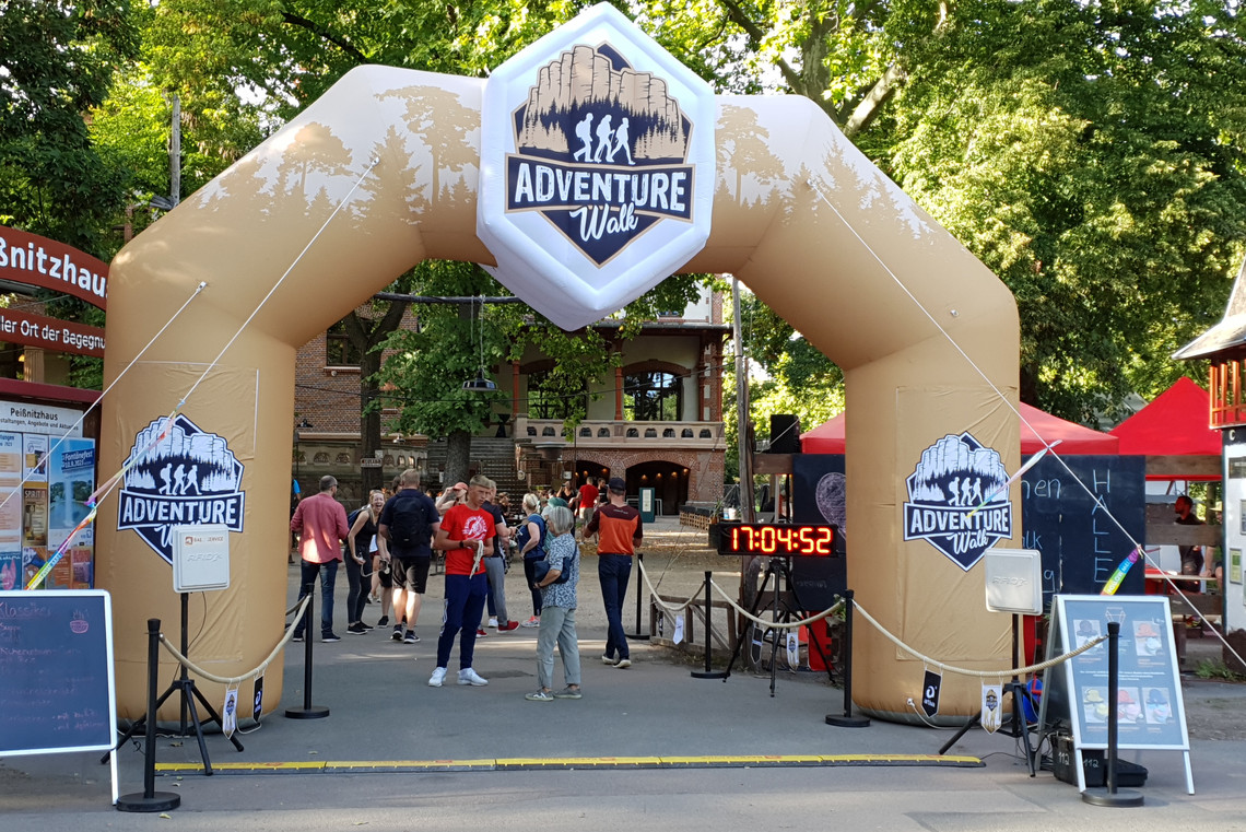 Aufblasbares Tor mit der Aufschrift „ADVENTURE Walk“ markiert den Eingang zu einer Veranstaltung im Freien, Menschen passieren die Absperrung, eine digitale Uhr zeigt 17:04:52.