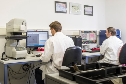 Zwei Fachkräfte in weißen Laborkitteln arbeiten an einem metallografischen Mikroskop und einem Computerarbeitsplatz in einem Prüflabor mit technischen Geräten und Monitoren.