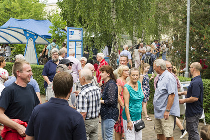 **Alt-Text:** Eine große Gruppe erwachsener Besucher\:innen unterhält sich bei einer Veranstaltung im Freien; im Hintergrund stehen Pavillons, Bäume und weitere Gäste auf einem belebten Weg.