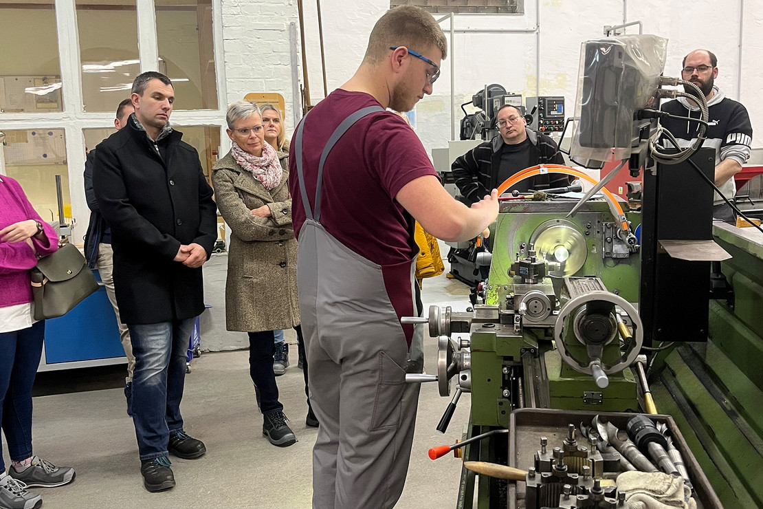 Eine Person arbeitet an einer Werkzeugmaschine und bearbeitet ein Metallbauteil, mehrere weitere Personen stehen dahinter und beobachten den Arbeitsprozess aufmerksam.