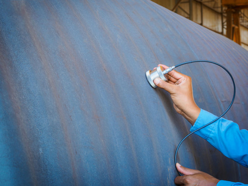 Hand legt einen Prüfkopf mit Kabel an eine große Metalloberfläche an, um die Materialdicke zu messen, im Hintergrund industrielle Umgebung.
