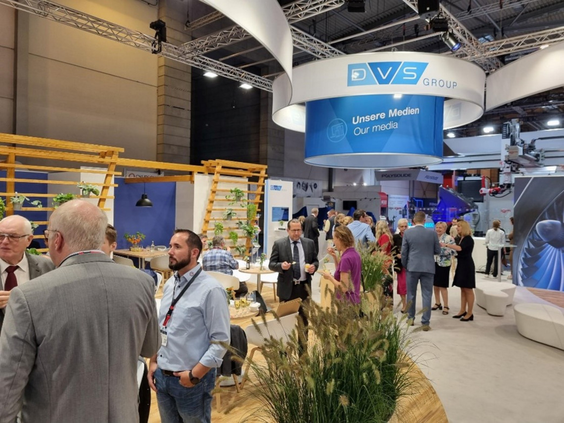 Besucherinnen und Besucher tauschen sich auf einem belebten Messestand der DVS Group aus; unter einem großen runden Banner mit der Aufschrift „Unsere Medien / Our media“ befinden sich Sitzbereiche mit Holz- und Pflanzenelementen.