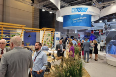 Besucherinnen und Besucher tauschen sich auf einem belebten Messestand der DVS Group aus; unter einem großen runden Banner mit der Aufschrift „Unsere Medien / Our media“ befinden sich Sitzbereiche mit Holz- und Pflanzenelementen.
