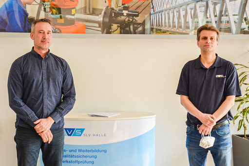 Zwei Männer stehen vor einem Infotresen der SLV Halle, links mit dunklem Hemd, rechts mit SLV-Poloshirt und Maske in der Hand, im Hintergrund Broschürenregal und Wandbild mit Industrieaufnahmen