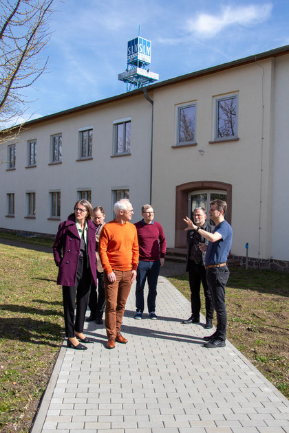 Fünf Männer und eine Frau stehen auf einem gepflasterten Weg vor einem hellen Gebäude mit dem Schriftzug „SLV Halle“ auf dem Dach, ein Mann erklärt etwas, die Gruppe blickt interessiert.