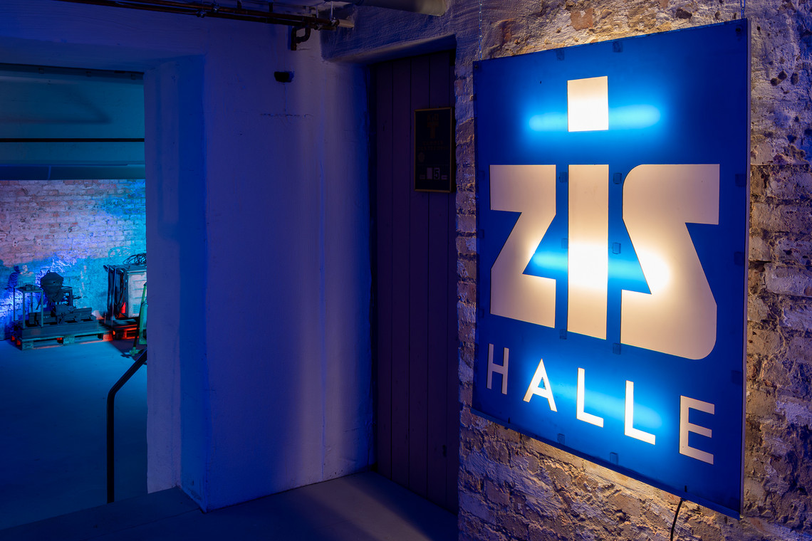 Leuchtendes ZIS-Halle-Schild an einer Backsteinwand, im Hintergrund blauer Lichtschein aus einem angrenzenden Raum