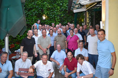 Gruppenfoto in sommerlicher Kleidung auf einer Terrasse mit Weinbewuchs im Hintergrund, einige sitzen auf Bänken, andere stehen dicht beieinander und lächeln in die Kamera.