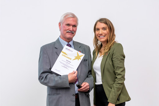 Zwei Personen stehen vor einem neutralen grauen Hintergrund nebeneinander, halten gemeinsam eine Urkunde und blicken in die Kamera.