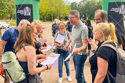 Vier Frauen und drei Männer stehen im Freien lachend in einer Gruppe zusammen und betrachten Urkunden. Im Hintergrund stehen grüne und schwarze Aufsteller zwischen Bäumen vom Firmenwandertag.
