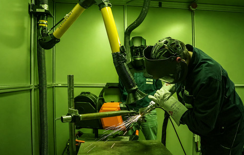 Eine Person mit Schweißhelm und Schutzkleidung beobachtet eine handgeführte Laserstrahlschweißarbeit, die in einer mit grünem Schutzglas ausgestatteten Kabine durchgeführt wird.