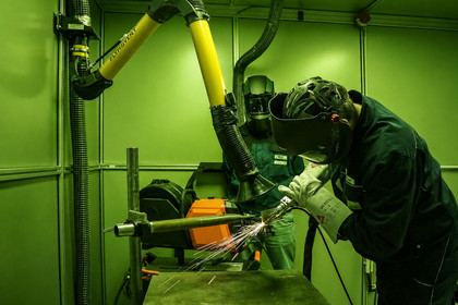 Eine Person mit Schweißhelm und Schutzkleidung beobachtet eine handgeführte Laserstrahlschweißarbeit, die in einer mit grünem Schutzglas ausgestatteten Kabine durchgeführt wird.