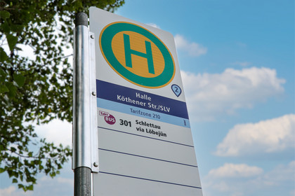 Ein Haltestellenschild mit grünem H auf gelbem Kreis steht vor blauem Himmel und Bäumen. Auf dem Schild sind der Haltestellenname und Angaben zum Linienverkehr zu sehen.