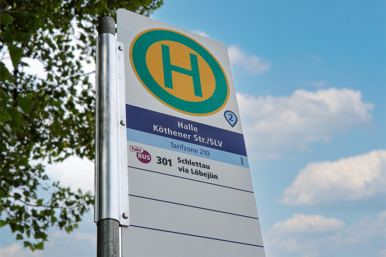 Ein Haltestellenschild mit grünem H auf gelbem Kreis steht vor blauem Himmel und Bäumen. Auf dem Schild sind der Haltestellenname und Angaben zum Linienverkehr zu sehen.