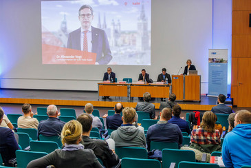 Personen sitzen in einem großen Vortragsraum, vorn auf der Bühne sprechen vier Personen an einem Pult, im Hintergrund ist eine Videoschaltung mit Oberbürgermeister Dr. Alexander Vogt projiziert.