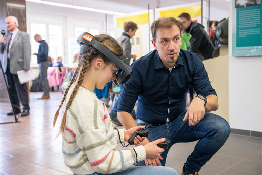 Ein Mann kniet neben einem sitzenden Mädchen mit geflochtenen Haaren und erklärt ihr die Benutzung einer VR-Brille, während im Hintergrund weitere Personen durch einen hellen Ausstellungsraum gehen.