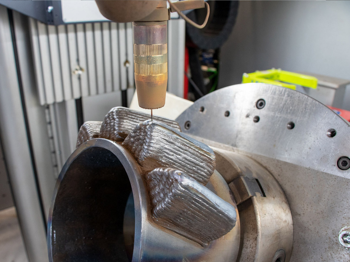 Nahaufnahme eines Metallbauteils mit mehreren aufgetragenen Schweißraupen. Darüber befindet sich eine Laserdüse im Prozess der additiven Materialauftragung.