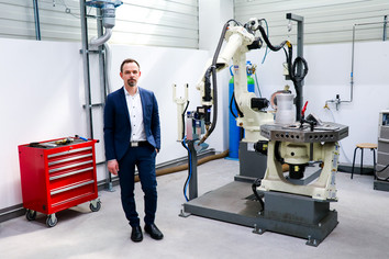 Mann im blauen Anzug steht in einer modernen Werkhalle neben einem weißen Industrieroboter mit Greifarm und einem rot lackierten Werkzeugwagen auf Rollen.