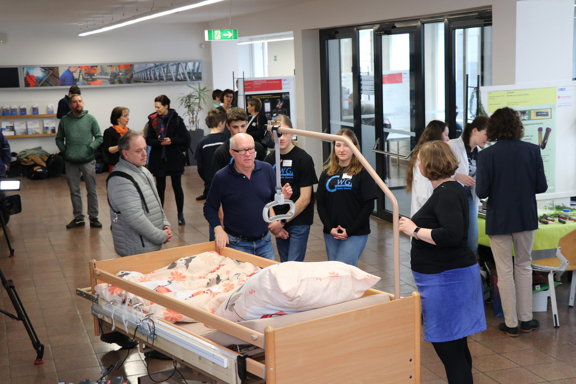  Jugendliche in schwarzen T-Shirts mit dem Logo „GWG“ präsentieren in elektrisch modifiziertes Pflegebett. Weitere Besuchende und Informationsstände sind im Hintergrund zu sehen.