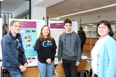 Drei junge Forschende präsentieren ihr Projekt an einem Stand mit wissenschaftlichen Postern. Zwei von ihnen tragen schwarze GWG-T-Shirts. Eine Frau im blauen Blazer steht mit ihnen im Gespräch.