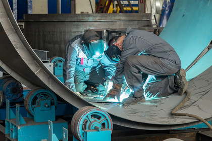 Ein junger Mann im karierten Hemd und blauer Latzhose prüft konzentriert mit einer Wasserwaage ein Metallteil, im Hintergrund arbeitet eine junge Frau an einem ähnlichen Werkstück.