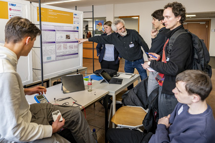 Ein Betreuungslehrer erklärt zwei Jury-Mitgliedern das Physik-Projekt seiner beiden Schüler. Auf dem Tisch befinden sich technische Geräte und Laptops.