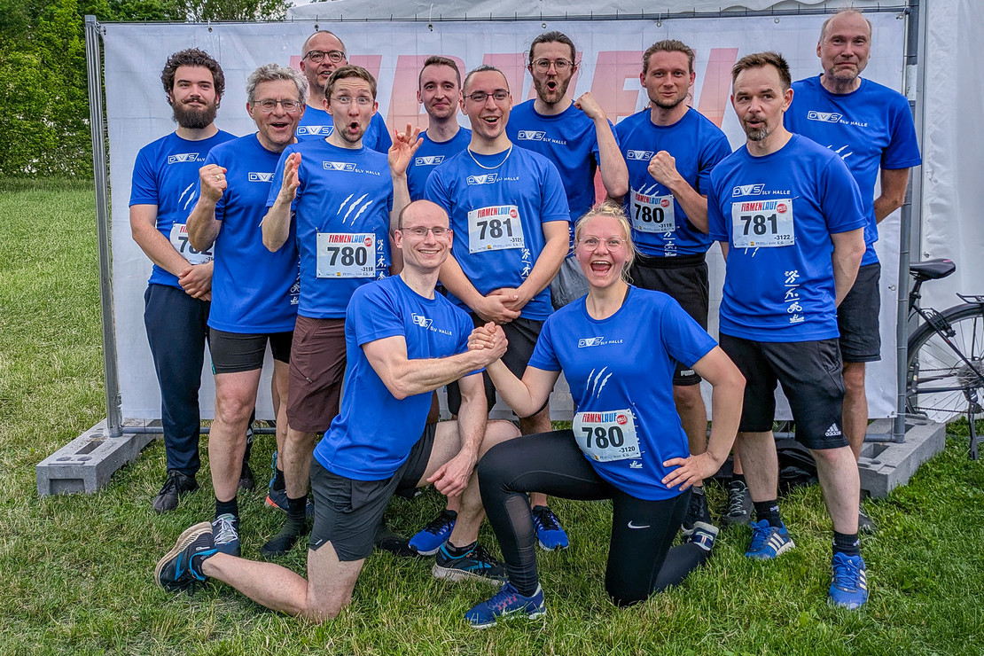 Gruppenfoto von elf Männern und einer Frau in blauen SLV Halle T-Shirts, sie posieren lachend und zeigen Jubelgesten vor einem Firmenlauf-Banner.