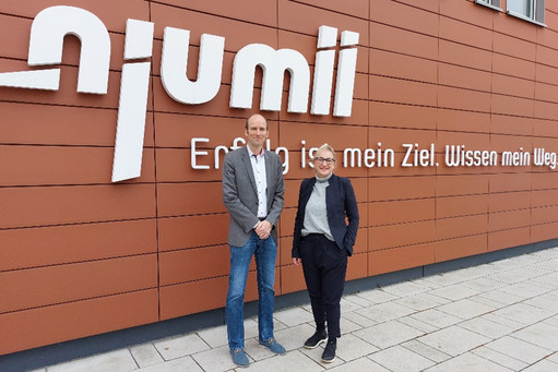 Ein Mann und eine Frau stehen lächelnd vor der Fassade des njumii-Gebäudes mit dem Schriftzug „Erfolg ist mein Ziel. Wissen mein Weg.“ 