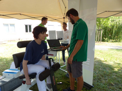 Eine Frau mit dunkelbraunen Haaren sitzt konzentriert an einem medizinischen Trainingsgerät unter einem Pavillon. Ein Mann in grünem Polohemd bedient ein Laptop, zwei weitere Männer stehen im Hintergrund neben einem Monitor.
