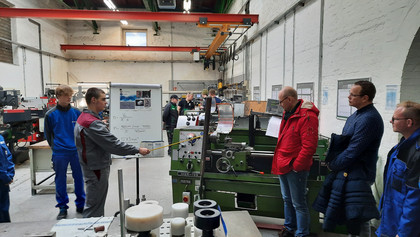 Mehrere Personen stehen in einer Werkhalle um eine Drehmaschine, eine Person im grauen Ausbildungsanzug erklärt technische Details an einem Whiteboard; im Hintergrund weitere Fachkräfte in blauer Arbeitskleidung.
