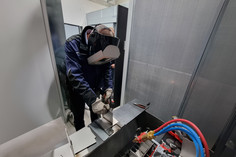 Eine Person mit Schweißschutzhelm und Handschuhen arbeitet konzentriert an einem Werkstück in einer Schweißkabine; im Vordergrund sind Gasanschlüsse mit farbigen Schläuchen sichtbar.