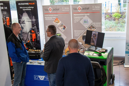 Drei Männer unterhalten sich an einem Messestand mit DVS-Tischdecke, im Hintergrund stehen Roll-ups der Marke VauQuadrat sowie ein Bildschirm mit technischem Inhalt; um sie herum sind Schweißgeräte und Prospektmaterialien platziert.