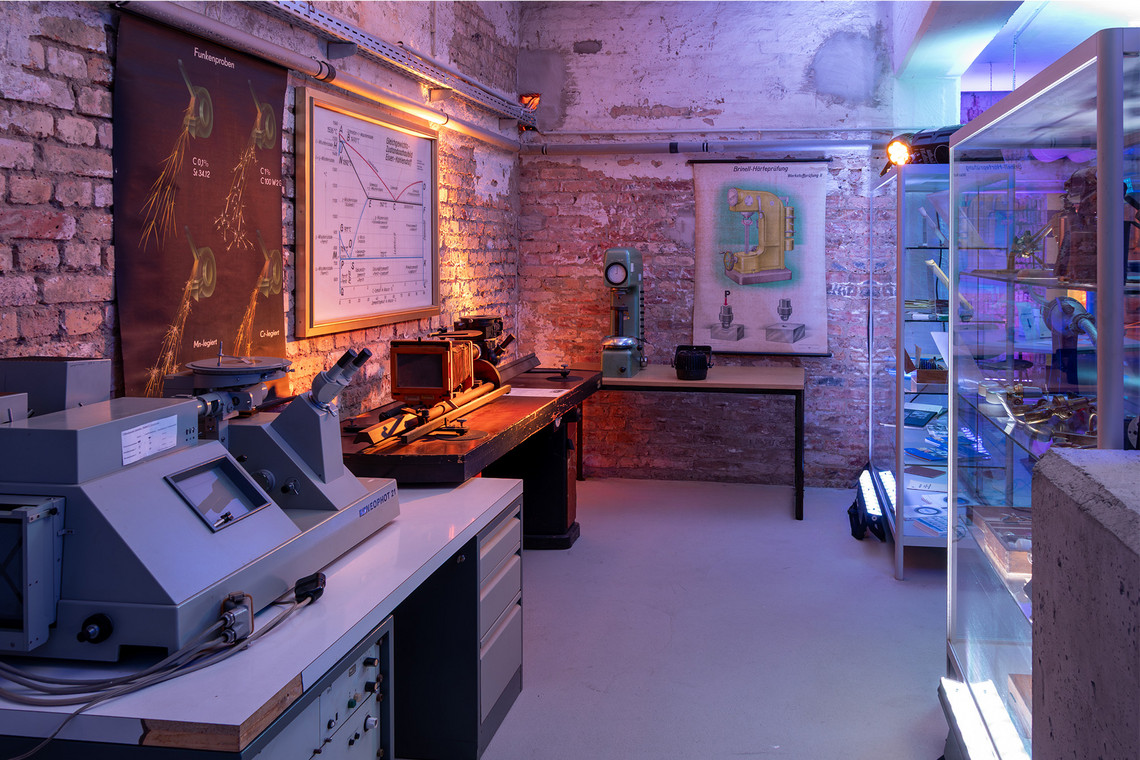 Ein Ausstellungsraum mit historischen Prüfgeräten, Mikroskop und Wandtafeln zur Werkstoffkunde zeigt die Entwicklung der Schweißtechnik in industrieller Umgebung.