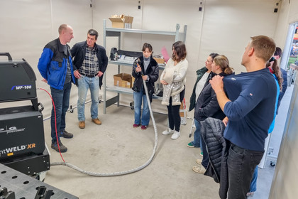 Mehrere Personen stehen in einer Werkstatt und hören einem Mann zu, der technische Geräte und Schweißtechnik erläutert.