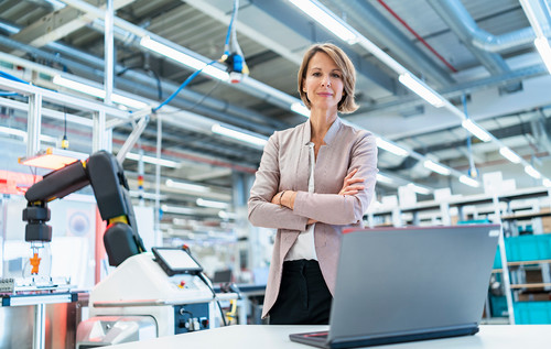 Frau mit überkreuzten Armen steht in einer Industriehalle mit Robotern vor einem Laptop.