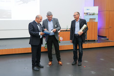 Drei Personen stehen auf einer Bühne und betrachten gemeinsam ein geöffnetes Präsentationsetui. Eine Person hält zusätzlich ein längliches Geschenk, im Hintergrund sind eine Projektionsfläche und ein Rednerpult mit dem Logo „DVS SLV Halle“ zu sehen.