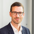 Dr. Rico Peters, im blauen Anzug mit Brille, lächelnd vor neutralem Hintergrund