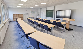 Großzügiger Tagungsraum geeignet für Präsentationen, mit Rednerpult, Beamer, Whiteboard, Tafel und leerem Regal für Materialien
