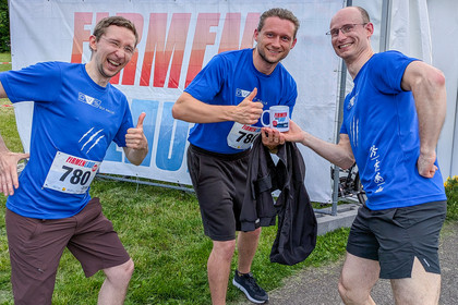 Drei Männer in blauen SLV Halle T-Shirts stehen lachend vor einem Firmenlauf-Banner, zeigen Daumen hoch und halten eine Tasse mit dem Logo des Firmenlaufs.
