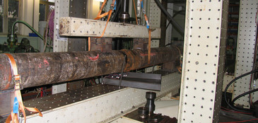 Ein großdimensionierter Holzprüfkörper mit Rissbildung ist in einer Schwingprüfmaschine eingespannt und wird über einen hydraulischen Zylinder belastet, um sein Tragverhalten zu analysieren.