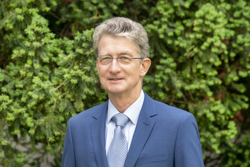 Ein Mann mit grauem Haar und Brille trägt einen blauen Anzug mit gestreifter Krawatte und steht vor einer grünen Hecke, freundlich lächelnd in die Kamera blickend.