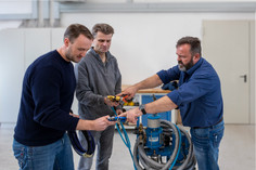 Drei Männer in Freizeitkleidung stehen in einer Werkhalle um ein technisches Gerät mit Schläuchen und analysieren gemeinsam eine Schlauchkupplung.