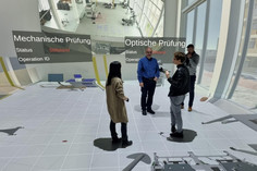 Eine Frau und drei Männer stehen in einem immersiven Raum mit Rundumprojektion, die eine digitale Laborumgebung mit Statusanzeigen für „Mechanische Prüfung“ und „Optische Prüfung“ simuliert; zwei Stationen zeigen den Status „Stillstand“.