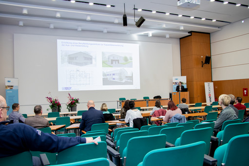 Teilnehmende sitzen in einem Konferenzraum mit grünen Stühlen, vorne läuft eine Präsentation über ein Erweiterungsprojekt, rechts steht ein Redner am Pult, Blumenarrangements auf dem Podium