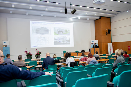 Teilnehmende sitzen in einem Konferenzraum mit grünen Stühlen, vorne läuft eine Präsentation über ein Erweiterungsprojekt, rechts steht ein Redner am Pult, Blumenarrangements auf dem Podium